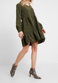 Femme portant une robe longue à manches longues en vert olive avec des volants et des bottines à talons en motif serpent sur un fond blanc uni.