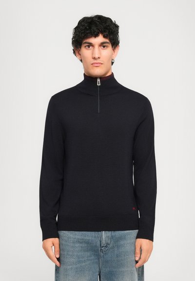 Man met krullend zwart haar die een zwarte sweater met halve ritssluiting en lichtblauwe spijkerbroek draagt, staande tegen een effen lichte achtergrond.