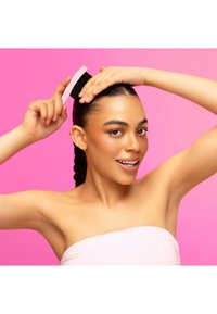 Czarna szczotka do włosów z różową rączką, trzymana przez osobę o opalonej cerze i piegach, na tle jasnoróżowego tła.