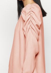 Dlouhá řasenková blůza z měkké broskvové látky, s řasením na ramenech a volným střihem. Hladká textura po celé ploše.