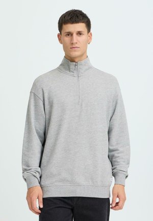 Junger Mann mit kurzem dunklem Haar, der ein hellgraues Sweatshirt mit Reißverschluss bis zur Brust und schwarze Hosen trägt, vor einem schlichten hellen Hintergrund stehend.