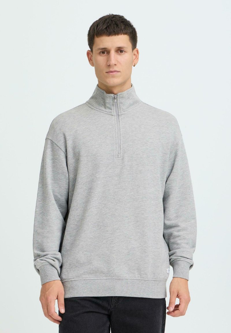 Jeune homme aux cheveux courts et foncés portant un sweat-shirt gris clair à demi-fermeture éclair et un pantalon noir, debout devant un fond clair uni.