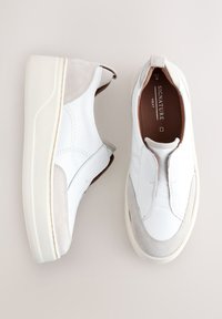 Zapatos de cuero blancos tipo slip-on con detalles en ante beige, cuello acolchado y suela blanca gruesa. El interior cuenta con forro marrón oscuro.