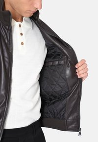 Giacca di pelle marrone aperta, che rivela un rivestimento imbottito nero. Presenta un colletto e polsini a coste, indossata sopra una polo bianca con texture.