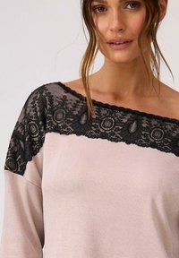 Długi rękaw bluzki w miękkim różowym materiale z czarną koronką na dekolcie, z okrągłymi wzorami i dekoracyjnymi akcentami.