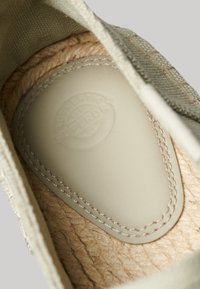 Interior de zapatilla de lona verde claro con plantilla texturizada beige que presenta un logotipo, bordes cosidos y material de yute natural por debajo.