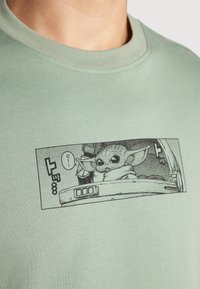 Ανοιχτοπράσινη βαμβακερή μπλούζα τύπου t-shirt με μαύρη εκτυπωμένη γραφιστική ενός χαρακτήρα με μεγάλα αυτιά σε κόμικ.