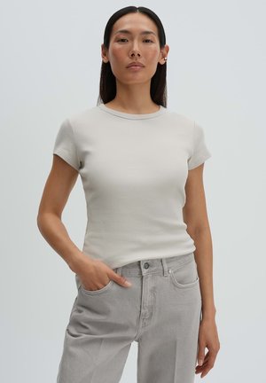 Donna con lunghi capelli scuri che indossa una maglietta a maniche corte a coste color grigio chiaro e jeans a vita alta color grigio chiaro, in piedi con una mano in tasca.