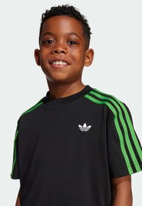 Svart t-shirt med gröna stänger på axlarna, korta ärmar och vit Adidas-logotyp på bröstet. Mjuk, texturerad tyg, avslappnad passform.