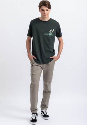 T-shirt in cotone verde scuro con una grafica di piedi nell'acqua, abbinato a pantaloni khaki chiari e sneakers bianco e nero.
