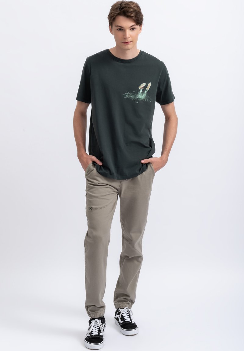 Donkergroene katoenen t-shirt met een afbeelding van voeten in water, gecombineerd met licht kaki broek en zwart-witte sneakers.