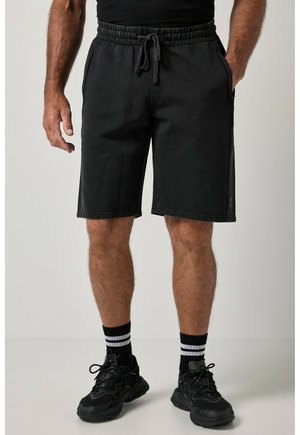 Mand iført sorte shorts med snøre, sorte sneakers og sorte sokker med hvide striber står mod en neutral baggrund.
