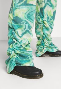 Calças com um padrão em verde e aqua em espiral, com detalhes franzidos nos tornozelos, combinadas com sapatos pretos de couro com costura amarela.