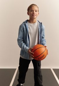 Veste en jean avec capuche sur un t-shirt blanc, tenant un ballon de basket orange. Un pantalon cargo foncé et des baskets noires complètent le look.