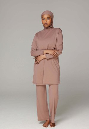 Woman wearing a modest, full-coverage mauve outfit with long sleeves, hijab, and wide-leg pants, standing barefoot against a plain background.