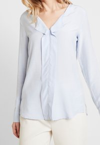 Blusa azzurra a maniche lunghe con scollo a V e pattina frontale sagomata. Texture liscia con un leggero lucido, orlo dritto.