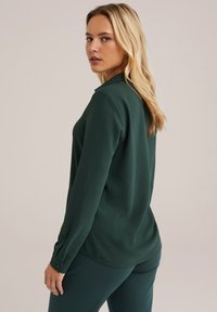 Žena s dlouhými blond vlasy, oblečená v tmavě zeleném tričku s dlouhými rukávy a odpovídajících kalhotách, se dívá přes rameno na neutrálním pozadí.