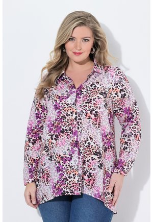 Frau mit langen blonden Haaren, die eine langärmelige, mehrfarbige lila- und pinke Leopardenmuster-Bluse und blaue Jeans trägt, steht vor einem weißen Hintergrund.