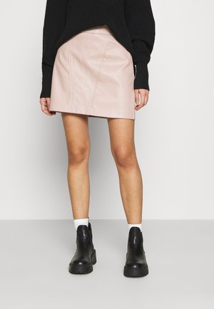 Vrouw met een lichtroze leren minirok, zwarte chunky enkellaarsjes, witte sokken en een zwarte gebreide trui, staand tegen een effen achtergrond.