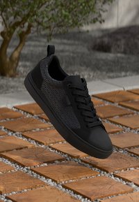 Adidași negri cu un brâu texturat, prezentând un model repetitiv al logo-ului, design cu șireturi și o talpă din cauciuc netedă, așezați pe o suprafață din piatră.