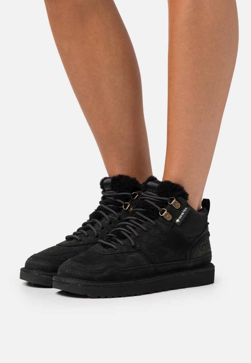 Bottes d'hiver noires mi-hautes avec doublure polaire, étiquette Gore-Tex, œillets métalliques et semelles épaisses en caoutchouc portées sur jambes nues.