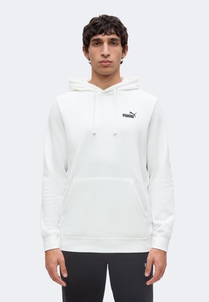 Man staat recht vooruit, draagt een witte Puma-hoodie met buidelzak en trekkoorden en zwarte broek tegen een effen lichte achtergrond.