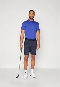 Blå poloskjorte, mørke shorts, svart belte, hvite sko. Står med en golfkølle, nøytral bakgrunn, fokus på antrekksdetaljer.