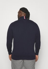 Pull en maille bleu foncé avec un col montant comportant une rayure rouge et blanche. Poignets et ourlets côtelés, fabriqué dans un tissu doux et texturé.