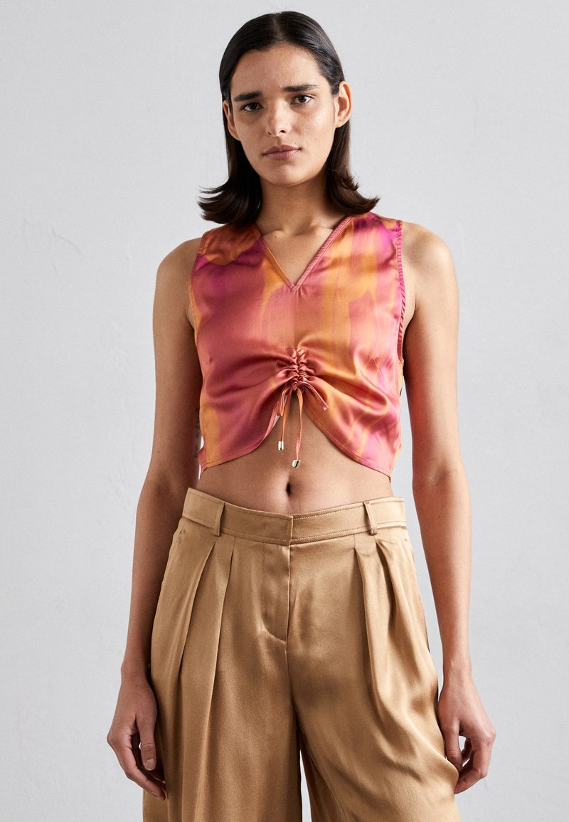 Kuttet topp i rosa og oransje gradient satin med V-hals og snor foran. Kombinert med høytlivs, beige bukser.