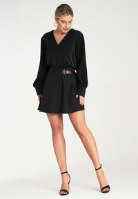 Czarna sukienka z dekoltem w kształcie litery V i długimi rękawami, z wcięciem w talii, ozdobiona brązowym paskiem. Spódnica jest rozkloszowana, sięgająca powyżej kolana.