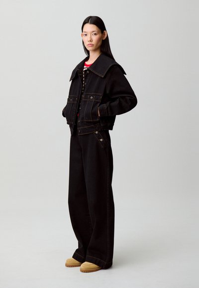 Donna in piedi con le mani nelle tasche, indossa pantaloni neri a gamba larga, giacca nera con grande colletto, camicia a righe e scarpe beige.