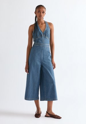 Kvinde med fletninger, iført en ærmeløs denim-dragt og brune flettede slingback-flats, stående foran en ensfarvet hvid baggrund.