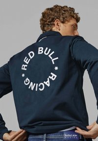 Mornarska jakna z dolgimi rokavi, ki na hrbtni strani prikazuje velik bel krog in napis "RED BULL RACING". Gladek material z klasičnim ovratnikom.