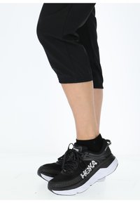 Sneaker atletici neri con suole bianche spesse, dotati di tomaie testurizzate e lacci neri. Indossati con pantaloni neri a metà polpaccio e calze nere.