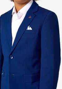 Tumši zila uzvalka žakete ar vienu pogu, iegrieztiem mēteļa lakatiem un kreiso krūšu kabatu, kurā ir balta un zila rakstaina kabatas kvadrāts.