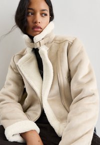 Beżowa kurtka z faux owczej skóry z wysokim kołnierzem, zapięciem na zamek błyskawiczny i kontrastowym białym podszyciem. Posiada boczną kieszeń i mocne szwy.