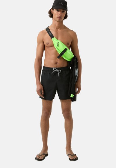 Shorts de bain noirs avec un cordon de serrage, associés à un sac à bandoulière vert néon vif. Porte également des tongs noires et une casquette noire.