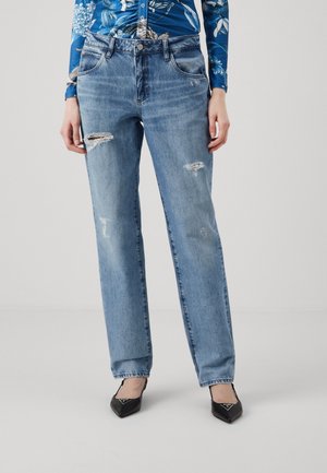 Persoon draagt lichtblauwe rechte spijkerbroek met slijtageplekken, een blauw gebloemd shirt met lange mouwen en zwarte pumps met puntige neus.