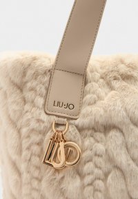 Sac fourre-tout beige en fausse fourrure avec une sangle en cuir lisse, arborant un logo doré et un charm avec les initiales décoratives. Design de surface texturé.