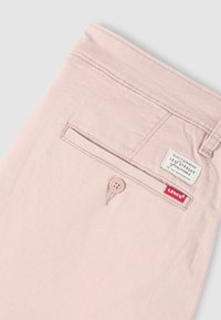 Ανοιχτό ροζ παντελόνια Levi's που δείχνουν την πίσω τσέπη με κούμπωμα κουμπιού, θηλιά για ζώνη και ετικέτες Levi's σε άσπρο και κόκκινο.