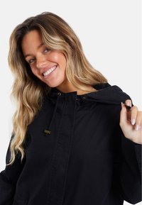Veste à capuche noire en tissu durable. Présente une fermeture à cordon de serrage et des accents en bronze. Texture lisse et coupe décontractée.