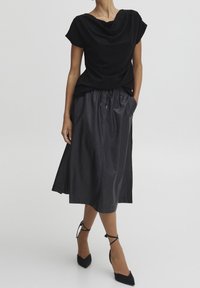 Zwarte gedrapeerde top met een ronde halslijn, gecombineerd met een zwarte, knielange rok met elastische taille. Model draagt puntige zwarte hakken.