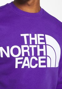 Fialová mikina s velkým bílým potiskem "THE NORTH FACE" a výrazným půlkruhovým logem. Hladká textura, žebrovaný límec.