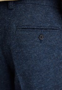 Pantaloni in misto lana blu scuro con motivo a spina di pesce, dotati di una tasca posteriore singola chiusa da un bottone nero.