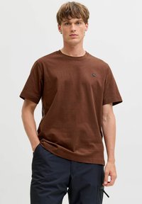 T-shirt en coton marron à manches courtes et col ras du cou, arborant un petit logo noir sur la poitrine gauche et une coupe décontractée.