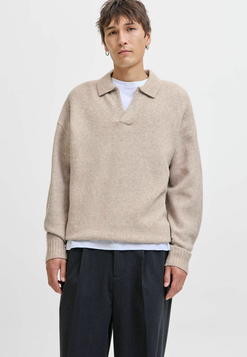 Lys beige sweater med krave, v-hals og ribbede manchetter, lavet af blødt stof. Båret over en hvid skjorte, parret med mørke bukser.