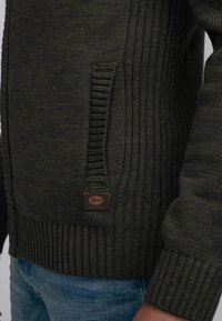 Mörkgrön stickad tröja med ribbad struktur, sidoficka och en liten läderetikett på nedre kanten. Långa ärmar med ribbade mudd.