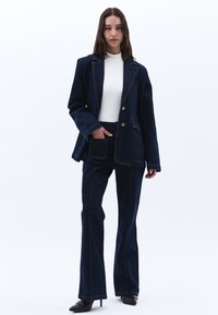 Tmavě modrý džínový blazer se shodnými kalhotami s rozšířenými nohavicemi. Mezi významné rysy patří kontrastní prošití a klasický design klopy.