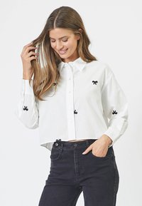 Camicia di cotone bianca con ricamo di fiocchi neri, chiusura a bottoni e orlo corto. Abbinata a jeans neri a vita alta.