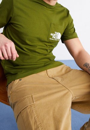 Mann trägt olivgrünes The North Face T-Shirt und hellbraune Cargo-Hose, sitzt mit teilweise sichtbarem Tattoo auf dem Unterarm.